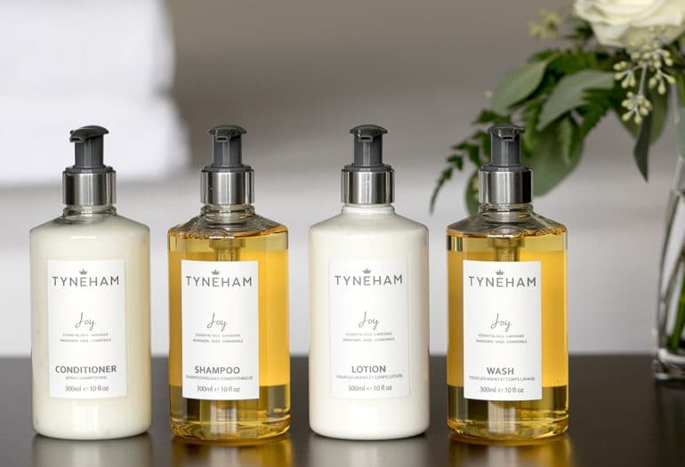 Tyneham conditioner, shampoo, lotion and body wash bottles with a vase of flowers in background