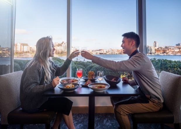 couple cheersing glasses with a variety of dishes on the table, a view of the harbour as the background
