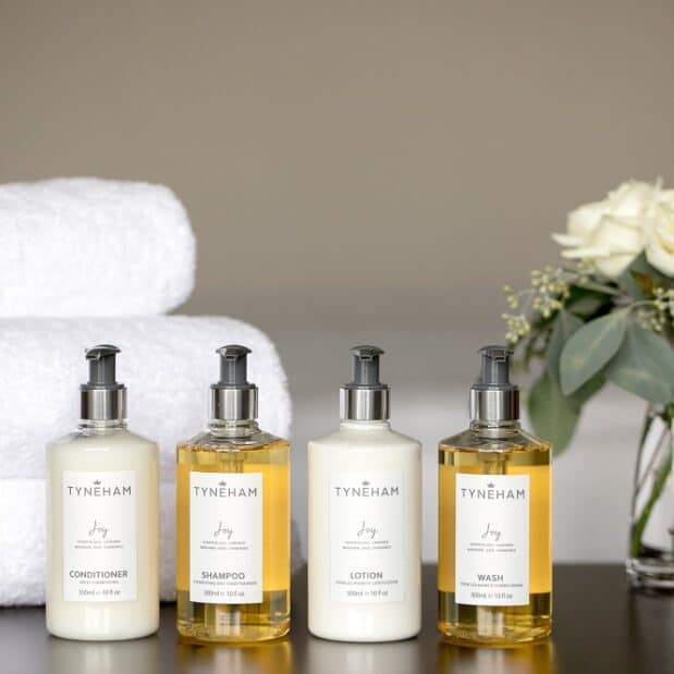 Tyneham conditioner, shampoo, lotion and wash bottles on a table with 3 stacked towels and a flower vase with white roses behind them
