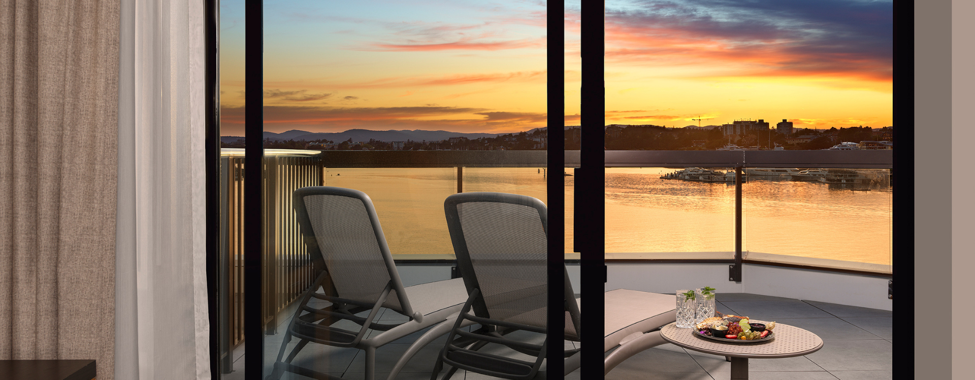 view of the sunset from the Laurel Wing sunset guestroom, 2 lounge chairs are seen from an open sliding door with a side table holding drinks and charcuterie on the balcony